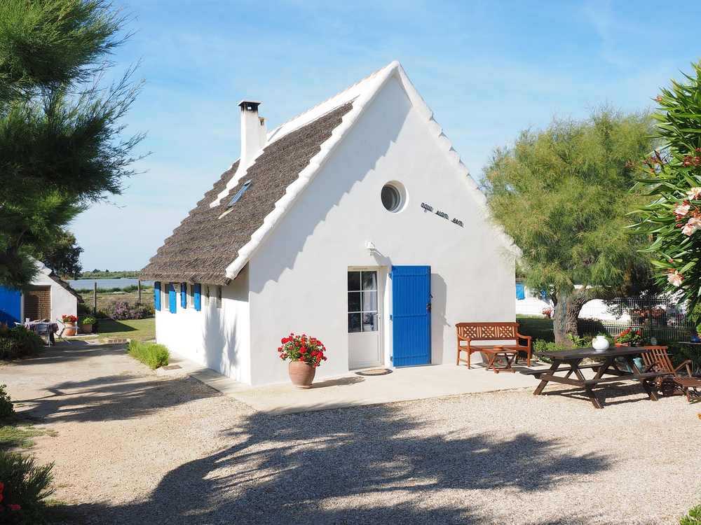 Maison blanche avec toit en chaume et porte bleue, entourée de jardin et de mobilier extérieur, sous un ciel bleu